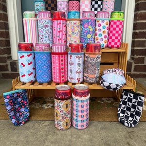 May include: A collection of colorful can coolers with various patterns, including cherries, stripes, floral designs, and smiley faces. The coolers are displayed on a wooden shelf and the ground. Some coolers have cans inside.