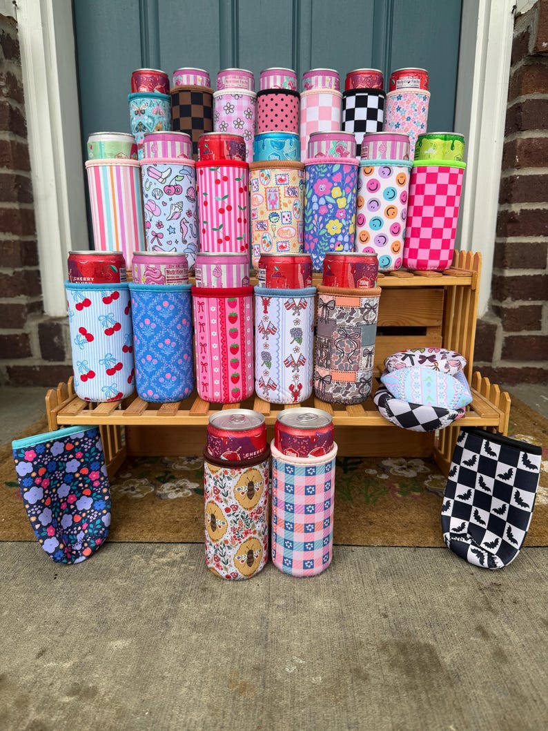 May include: A collection of colorful can coolers with various patterns, including cherries, stripes, floral designs, and smiley faces. The coolers are displayed on a wooden shelf and the ground. Some coolers have cans inside.