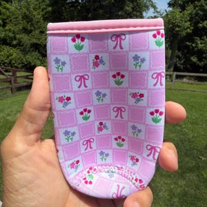 May include: Pink and white patterned beverage holder with a floral and bow design. The holder is made of a soft, flexible material and has a pink trim around the top. The design includes squares with flowers and bows.