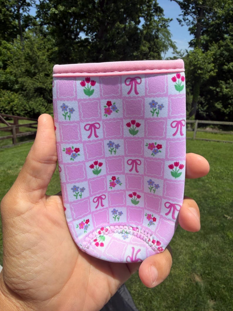 May include: Pink and white patterned beverage holder with a floral and bow design. The holder is made of a soft, flexible material and has a pink trim around the top. The design includes squares with flowers and bows.