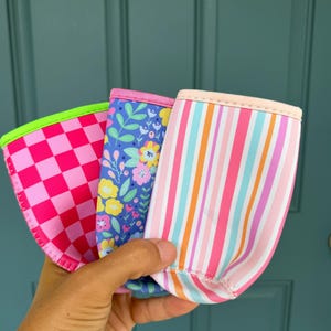 May include: Three colorful beverage insulators. One has a pink and red checkerboard pattern, another has a blue floral design, and the third has pink and orange stripes. Each has a contrasting trim.