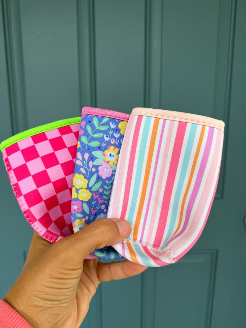 May include: Three colorful beverage insulators. One has a pink and red checkerboard pattern, another has a blue floral design, and the third has pink and orange stripes. Each has a contrasting trim.