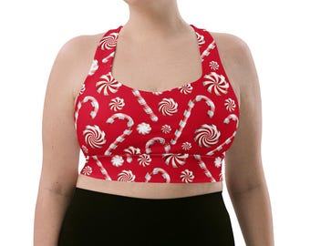 May include: A red racerback sports bra featuring a repeating pattern of white and red candy canes and peppermint swirl candies. The garment has a scoop neckline and a cropped hemline, providing a fitted silhouette with a festive holiday print.