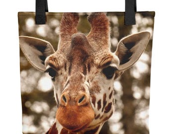 May include: A tote bag featuring a high-resolution, close-up photograph of a giraffe's face. The giraffe has brown and tan patterned fur, large dark eyes, and prominent ears. The bag includes two sturdy black fabric handles at the top.