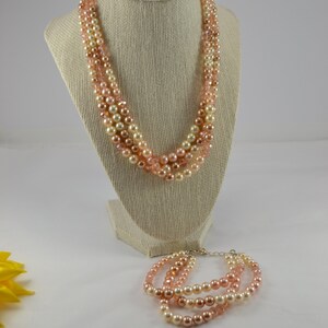 May include: A multi-strand necklace and bracelet set made of pink, peach, and cream coloured pearls. The necklace is displayed on a mannequin bust and the bracelet is lying on a white surface.
