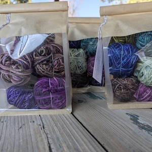 May include: A set of six skeins of yarn in various shades of purple, blue, and green. The yarn is packaged in a clear plastic bag with a brown paper label that says "The Yarn Harlot -  Shrug Yarn".