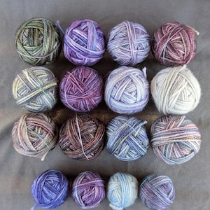 May include: An overhead shot of 16 balls of yarn in various colors, including purple, blue, green, and brown. The yarn balls are arranged in four rows of four. Each ball is tightly wound, and some have a rubber band around them.