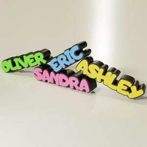 Puede incluir: Cuatro letreros con nombres en 3D con una fuente l&uacute;dica. Los nombres son "Oliver" en verde, "Eric" en azul, "Sandra" en rosa y "Ashley" en amarillo. Cada nombre est&aacute; sobre un fondo negro.