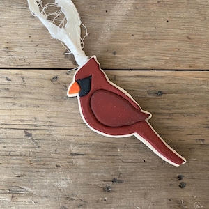 May include: A red cardinal bird ornament with a white ribbon. The ornament is made of ceramic and has a painted finish.
