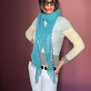 Könnte beinhalten: Eine Frau trägt einen hellblauen, weißen und beige gestreiften Pullover mit einem hellblauen Schal. Sie trägt eine weiße Hose und einen schwarzen Gürtel. Sie trägt auch eine Sonnenbrille.