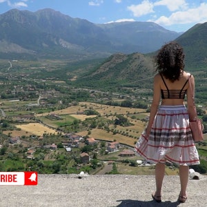 May include: A woman in a black crop top and a plaid skirt stands on a cliff overlooking a valley with rolling hills and a small town in the distance. The sky is blue with white clouds.