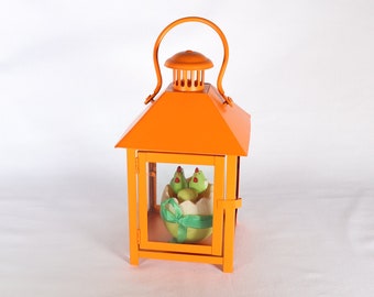 May include: Orange metal lantern with a glass window, featuring a white egg with two green chicks inside. The lantern has a handle on top.