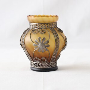 May include: A small, amber glass vase with a silver metal design. The vase has a floral pattern and a geometric border.