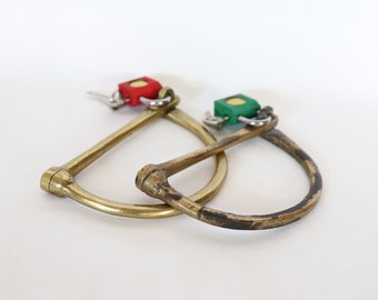 Puede incluir: Dos anillos de latón en forma de D con un candado rojo y verde unido a cada uno. Los candados están cerrados y los anillos están entrelazados.