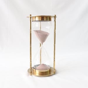 Puede incluir: Un reloj de arena de latón con un bulbo de vidrio transparente y arena rosa. El reloj de arena está sobre una superficie blanca.
