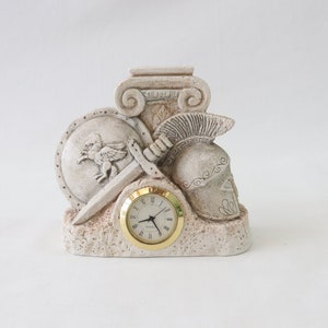 May include: A white ceramic clock with a Roman numeral face, featuring a sculpted design of a sword, shield, helmet, and column.