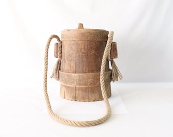 Puede incluir: Un cubo de madera con un asa de cuerda y borlas. El cubo está hecho de madera natural y tiene un aspecto rústico.
