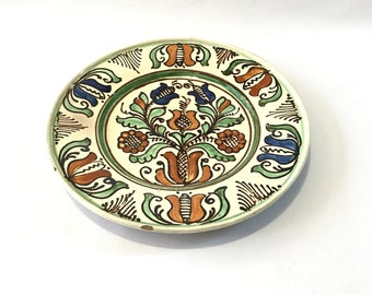 May include: A white ceramic plate with a green, blue, and orange floral design. The plate has a white background with a green border and a floral design in the center.