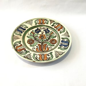 May include: A white ceramic plate with a green, blue, and orange floral design. The plate has a white background with a green border and a floral design in the center.
