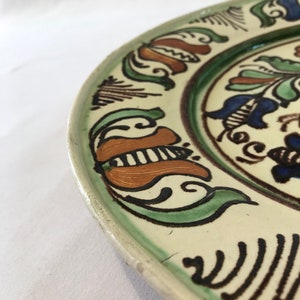May include: A white ceramic plate with a green, brown, and blue floral design. The plate has a raised rim and a slightly scalloped edge.