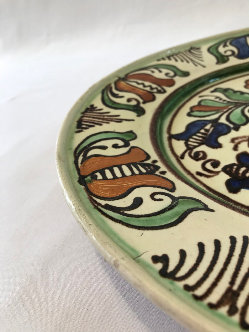 May include: A white ceramic plate with a green, brown, and blue floral design. The plate has a raised rim and a slightly scalloped edge.