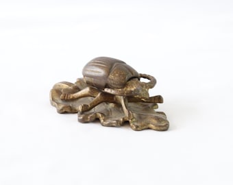 May include: A gold-toned metal beetle figurine with a hinged lid, sitting on a leaf-shaped base. The beetle is detailed with textured wings and legs.