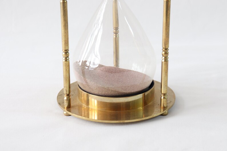 May include: A brass hourglass with a clear glass bulb and a round base. The sand is a light brown color.