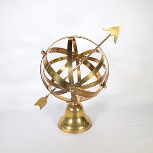 May include: A gold-colored brass armillary sphere with an arrow pointing upwards. The sphere is on a round base.