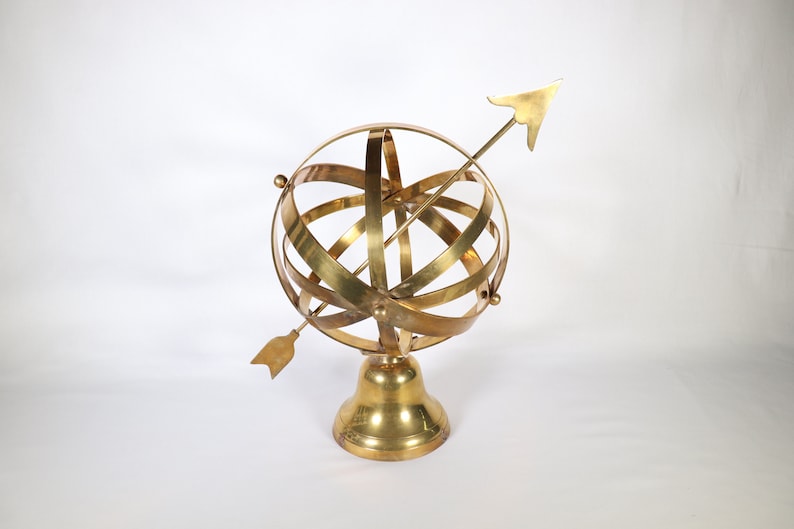 May include: A gold-colored brass armillary sphere with an arrow pointing upwards. The sphere is on a round base.