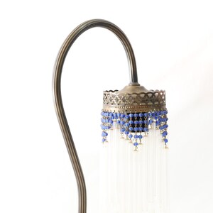 K&ouml;nnte beinhalten: Eine Stehlampe aus Messing mit einem gebogenen Arm und einem dekorativen Schirm. Der Schirm ist mit blauen Perlen und durchsichtigen Glasf&auml;den verziert.
