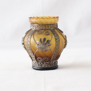 May include: A small, amber glass vase with a decorative metal design. The vase has a scalloped rim and is decorated with silver-colored metal flowers and geometric patterns.