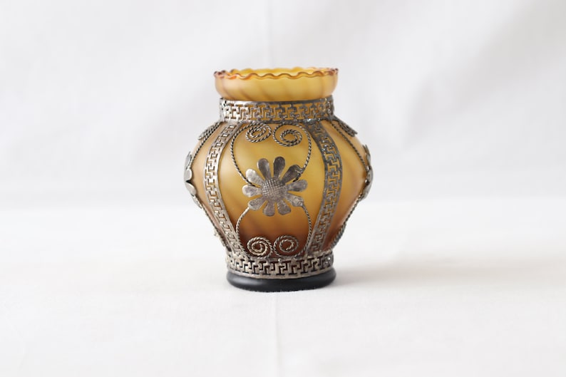 May include: A small, amber glass vase with a decorative metal design. The vase has a scalloped rim and is decorated with silver-colored metal flowers and geometric patterns.