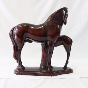 May include: A dark brown ceramic sculpture of a horse and foal standing side by side. The horse is standing with its head turned to the side, while the foal is standing with its head lowered. The sculpture is set on a rectangular base.