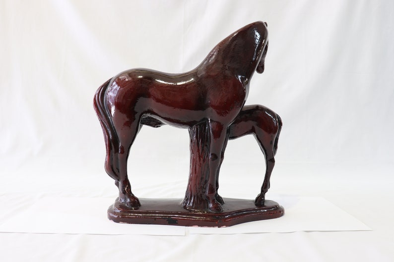 May include: A dark brown ceramic sculpture of a horse and foal standing side by side. The horse is standing with its head turned to the side, while the foal is standing with its head lowered. The sculpture is set on a rectangular base.