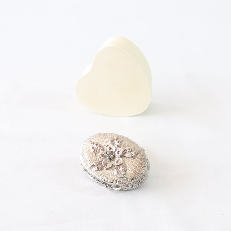 May include: A silver oval trinket box with a floral design featuring pink rhinestones. The box is sitting on a white surface.