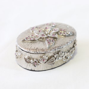 May include: A silver oval trinket box with a floral design. The lid features a large flower with pink rhinestones. The box is decorated with smaller flowers and leaves around the edge.