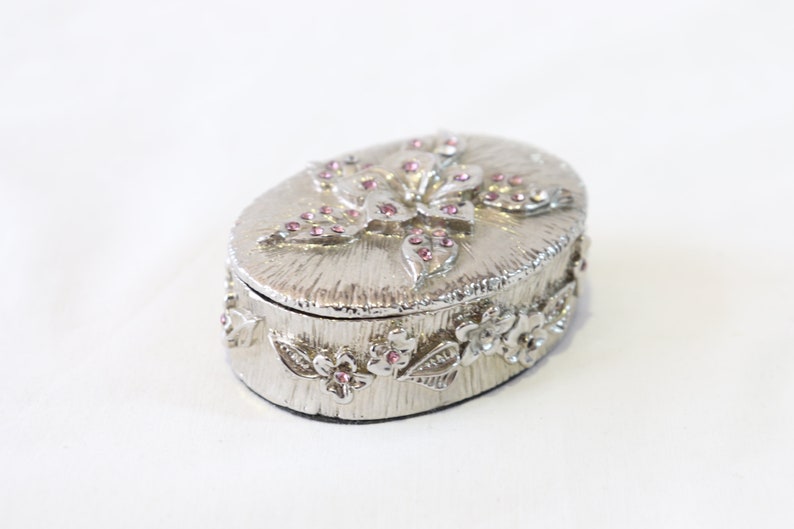 May include: A silver oval trinket box with a floral design. The lid features a large flower with pink rhinestones. The box is decorated with smaller flowers and leaves around the edge.