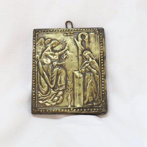 May include: A gold-colored metal plaque with a raised design depicting an angel and a woman. The angel is holding a branch with leaves and the woman is holding a book. The plaque has a decorative border.
