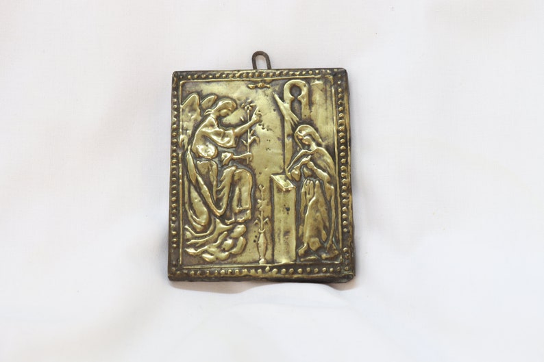 May include: A gold-colored metal plaque with a raised design depicting an angel and a woman. The angel is holding a branch with leaves and the woman is holding a book. The plaque has a decorative border.