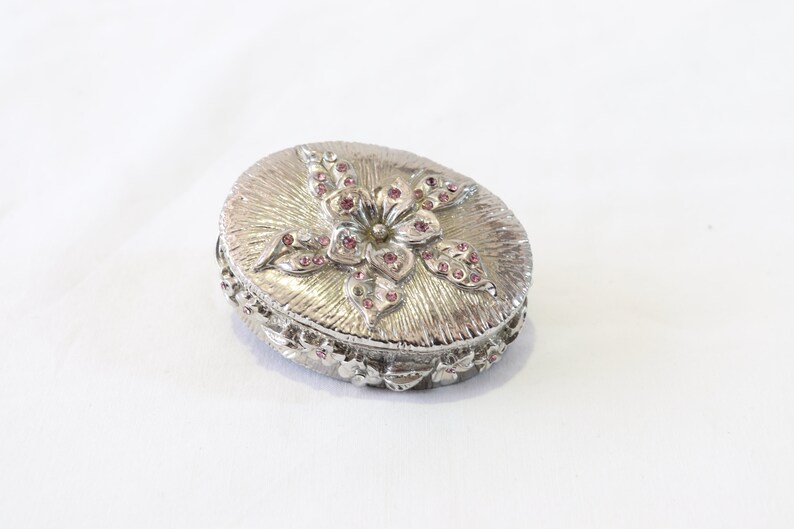 May include: A silver oval trinket box with a floral design. The flower is made of pink rhinestones and has a silver centre.
