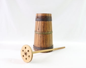 Könnte beinhalten: Eine Holzbutterfass mit einem Holzrührlöffel. Das Fass ist aus Holz und hat Metallbänder darum. Der Rührlöffel hat einen runden Holzgriff und einen flachen, runden Kopf mit Löchern.