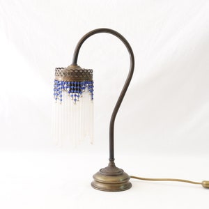 K&ouml;nnte beinhalten: Eine Vintage-Tischlampe aus Messing mit einem wei&szlig;en Glasschirm und einem blauen Perlenbesatz. Die Lampe hat einen flexiblen Arm und einen runden Sockel.