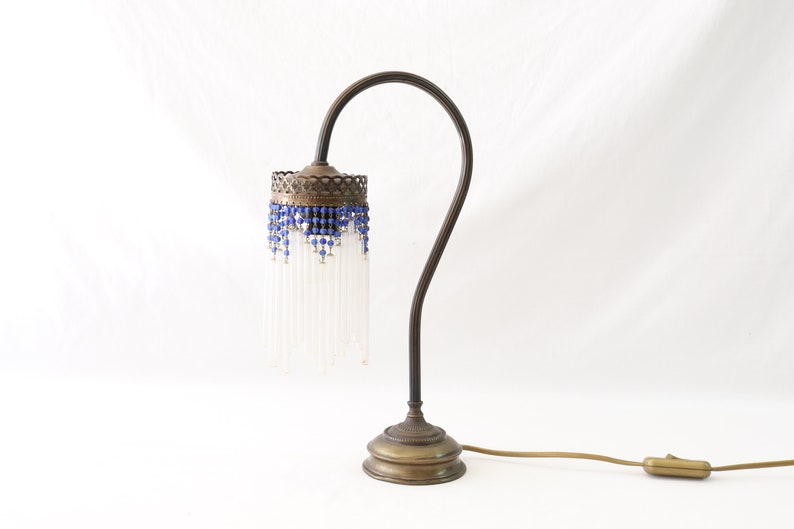 K&ouml;nnte beinhalten: Eine Vintage-Tischlampe aus Messing mit einem wei&szlig;en Glasschirm und einem blauen Perlenbesatz. Die Lampe hat einen flexiblen Arm und einen runden Sockel.