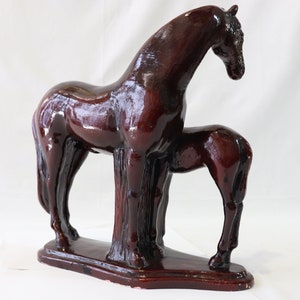May include: A dark brown ceramic sculpture of a horse and foal standing side by side. The horse is larger than the foal and has a long, flowing mane. The sculpture is on a rectangular base.
