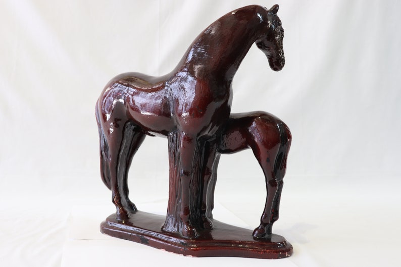 May include: A dark brown ceramic sculpture of a horse and foal standing side by side. The horse is larger than the foal and has a long, flowing mane. The sculpture is on a rectangular base.