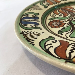 May include: A white ceramic plate with a green, brown, and blue floral design. The plate has a raised rim and a glossy finish.