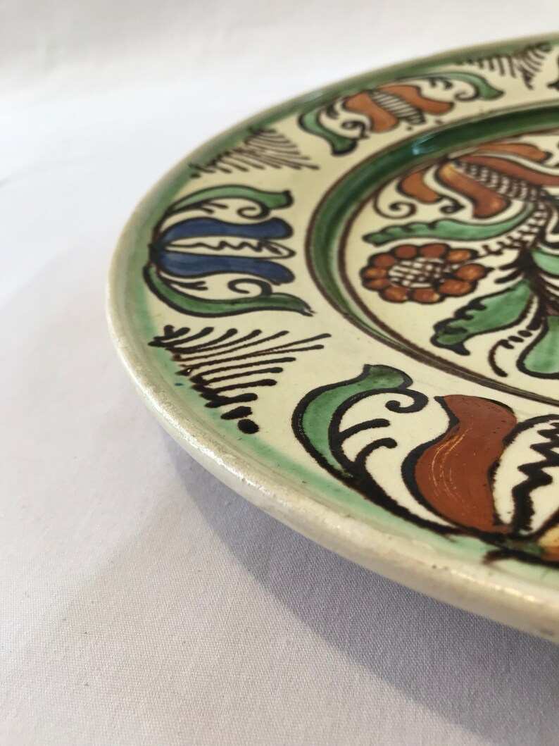 May include: A white ceramic plate with a green, brown, and blue floral design. The plate has a raised rim and a glossy finish.