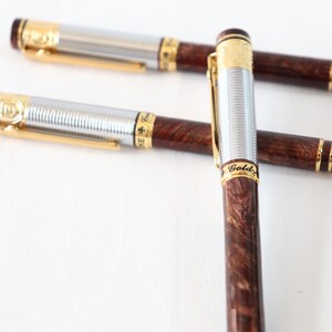 May include: Three gold and silver pens with brown wood-grain barrels. The pens have gold accents and the words "Gold" and "Swiss" are visible on the barrels.