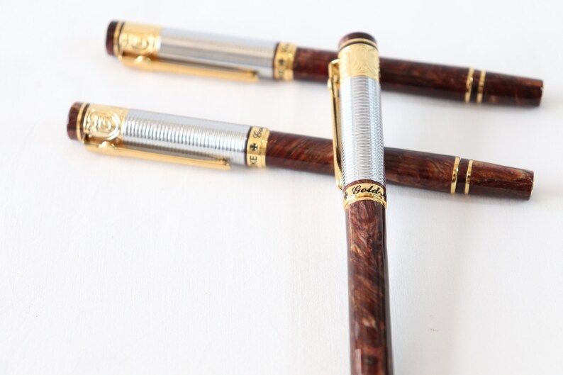 May include: Three gold and silver pens with brown wood-grain barrels. The pens have gold accents and the words "Gold" and "Swiss" are visible on the barrels.