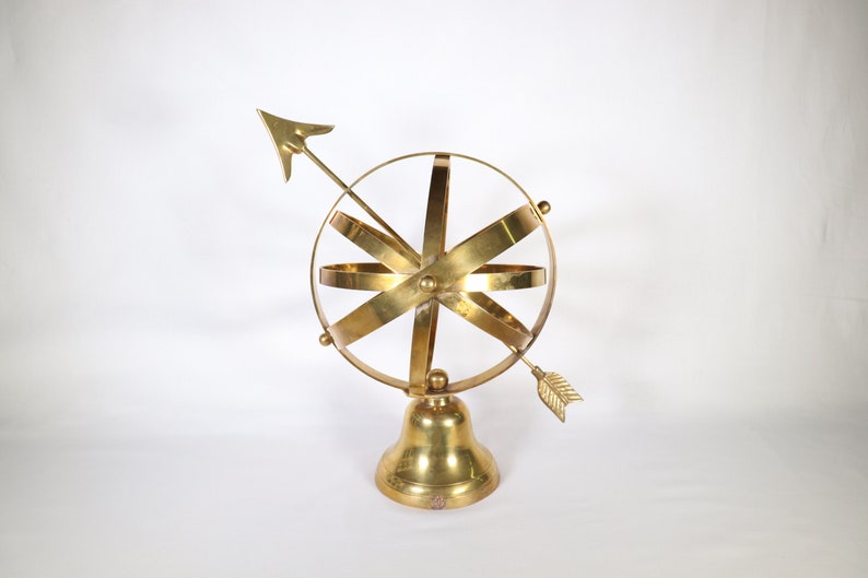 May include: A gold-colored metal sculpture of a sphere with a compass arrow pointing to the right. The sphere is on a bell-shaped base.